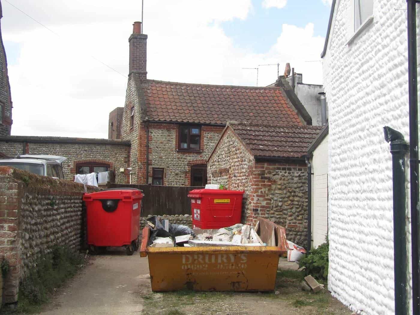 A small builders skip full of rubble in a narrow driveway between brick houses