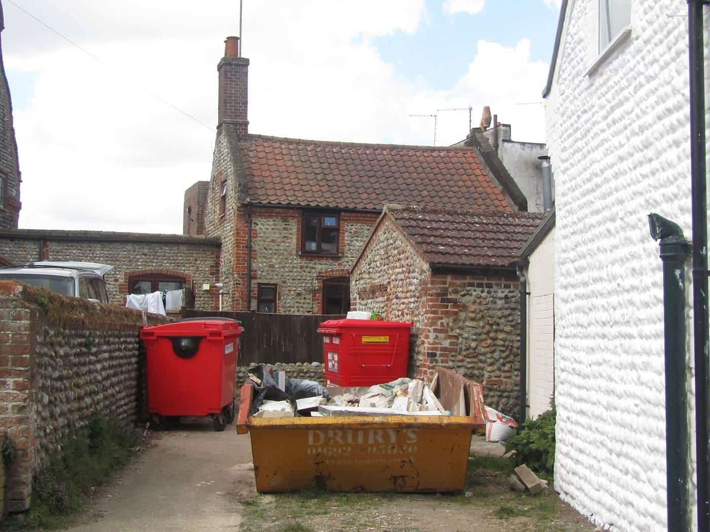 A small builders skip full of rubble in a narrow driveway between brick houses