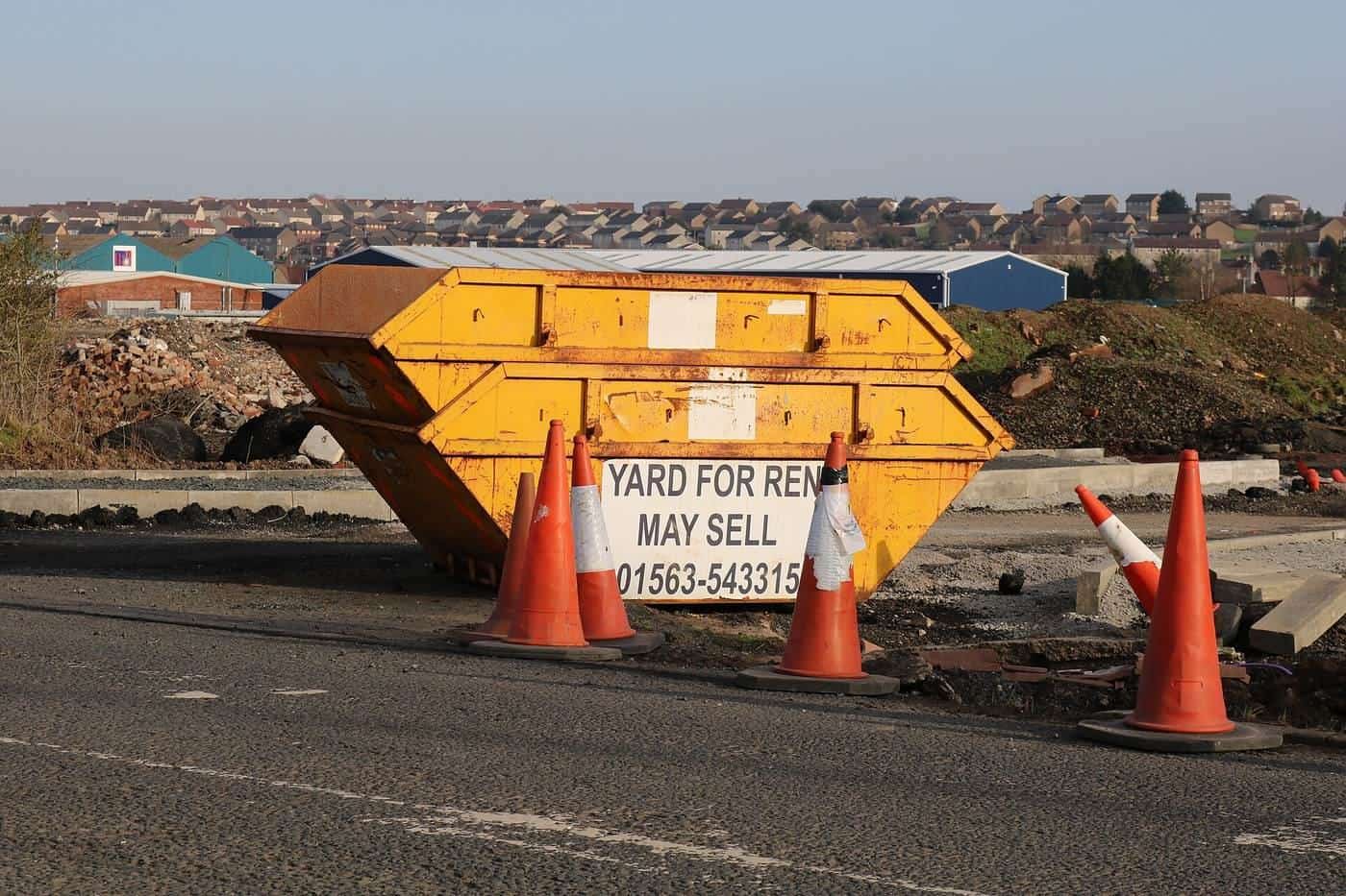 Two empty yellow skips stacked on a roadside with traffic cones